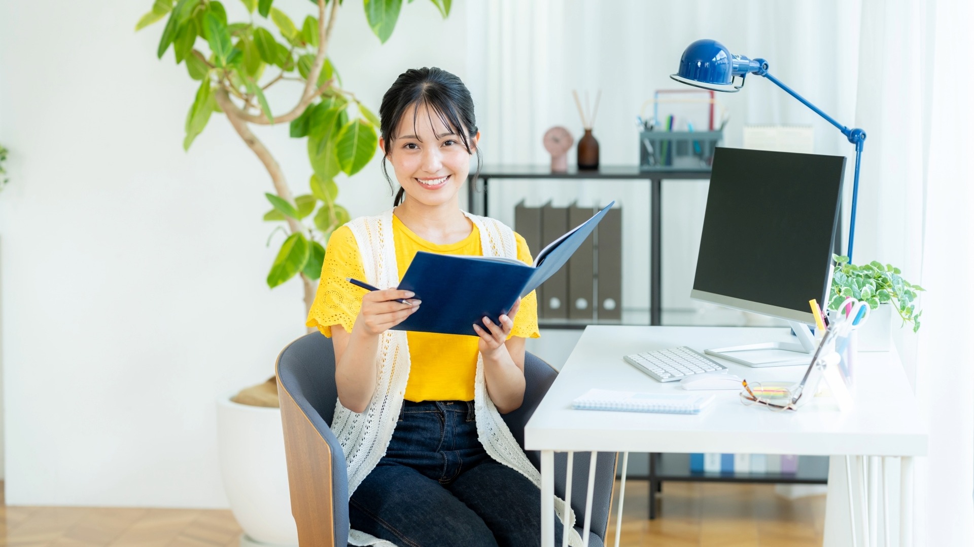 リモートワークでオウンドメディアのコンテンツ企画を練り、青いノートを手に微笑む女性。