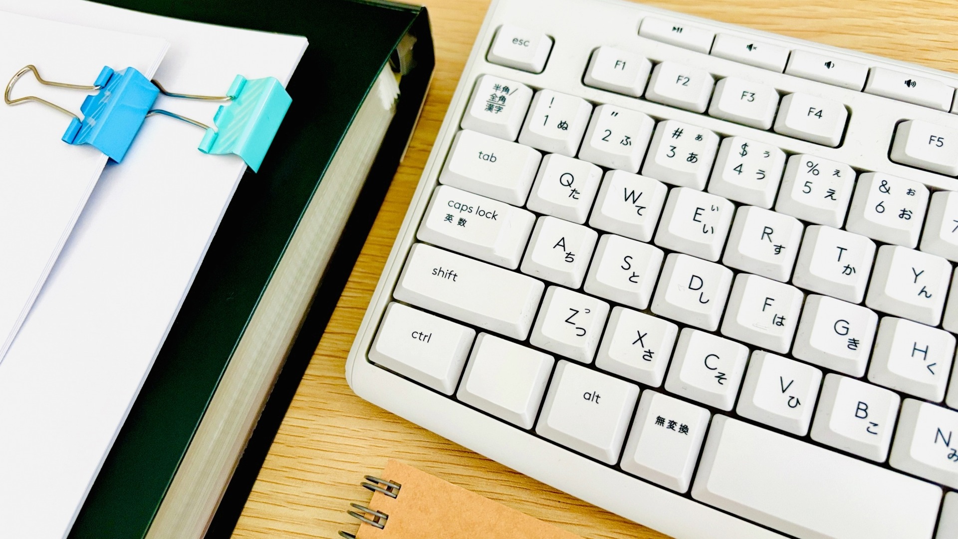 白いフルキーボードの横に、青い大きなクリップでまとめられた白い書類と、茶色のノート。